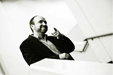 A man in a suit jacket and striped shirt is smiling and pointing forward with his right hand, standing indoors against a modern, angled background in a black-and-white photo.